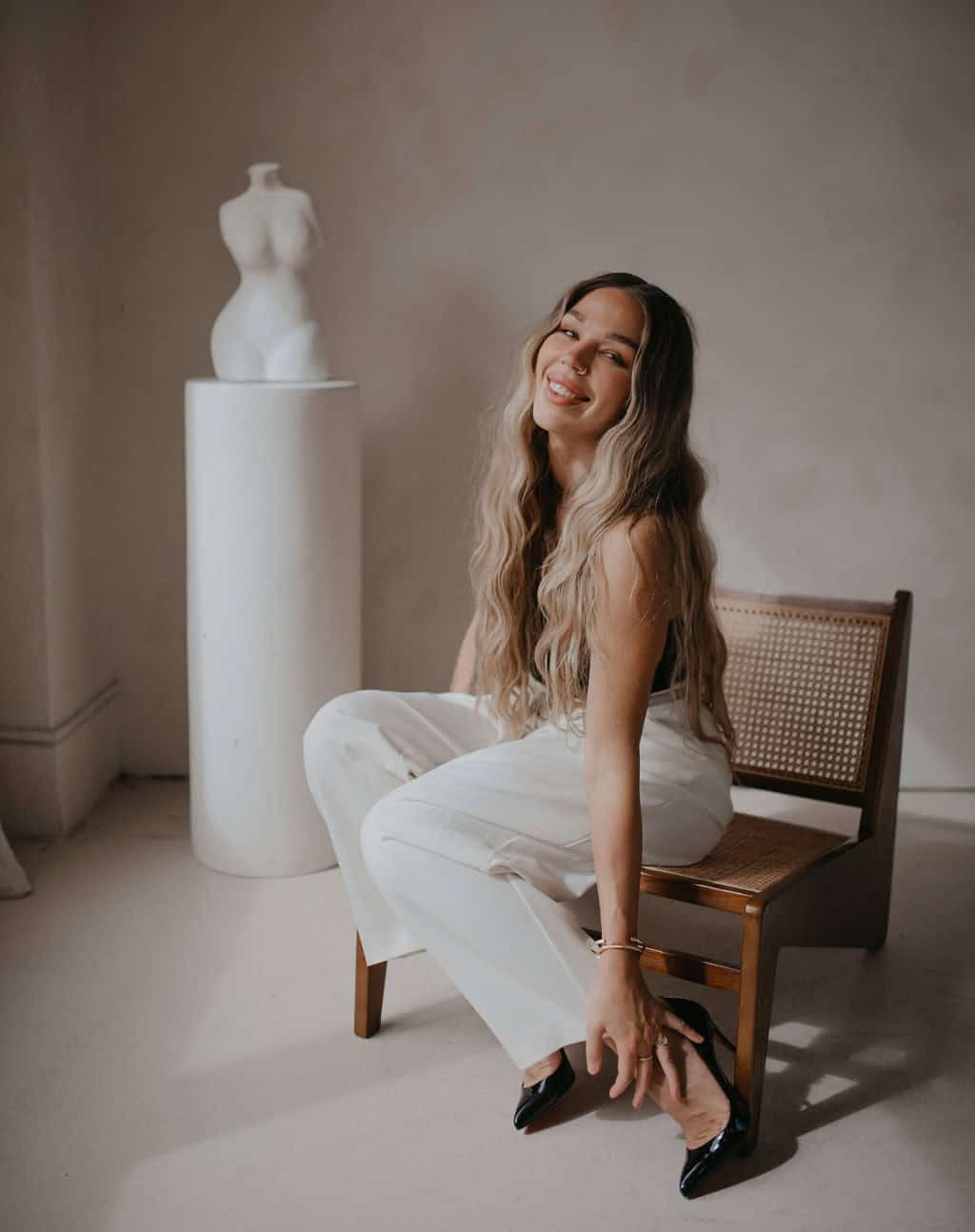 Elegant woman with long wavy hair smiling, sitting on a wooden chair in a minimalist studio setting, showcasing Jaime Marie’s fashion styling expertise in Chilliwack and Vancouver, BC.