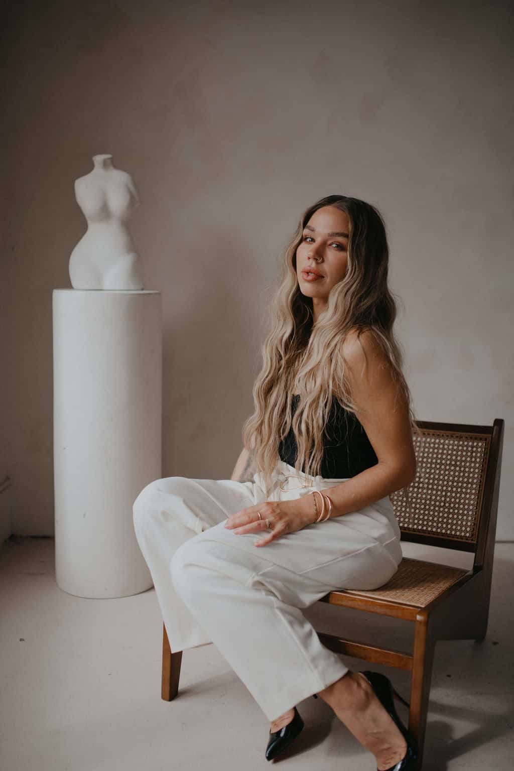 Elegant woman with long wavy blonde hair sitting on a wooden chair in a minimalistic studio space in Vancouver, BC, representing Jaime Marie’s artistry and creative style for her Chilliwack, BC, and online clients.